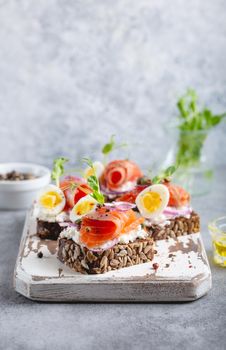 Close-up of sandwich with dark rye bread, cream cheese, salmon, onion, capers, boiled egg on white wooden cutting board, concrete background. Traditional Scandinavian open sandwich Smorrebrod.