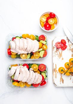 Healthy meal prep for lunch, diet boiled chicken fillet with vegetables salad in lunch boxes containers top view on white marble kitchen table background with knife and cutting board