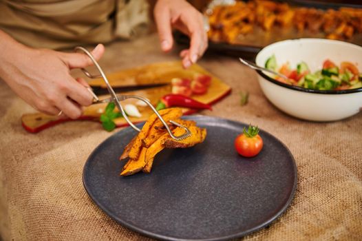 Close-up of serving dishes. A housewife standing by a kitchen table, puts baked slices of sweet potato on a serving plate. Delicious healthy vegan meal with wholesome dieting organic batata