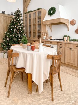 Modern, light, festive, cozy kitchen interior with Christmas and New Year decorations, kitchen table, utensils, copper pans on wall and big Christmas tree with presents, winter holidays concept