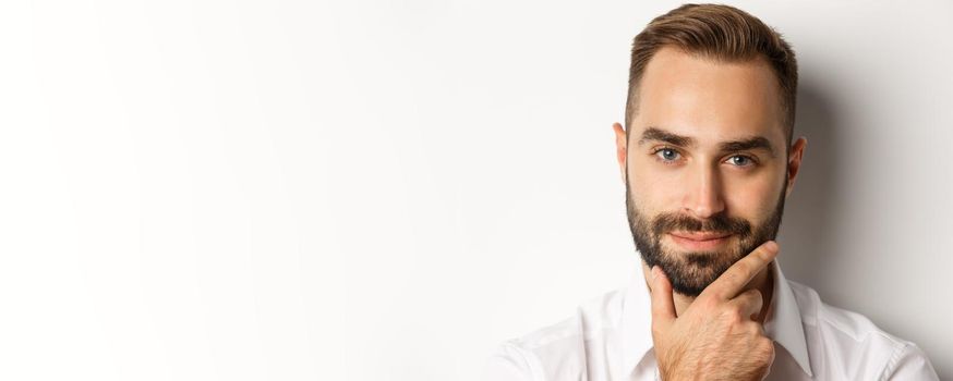 Emotions and people concept. Headshot of handsome thoughtful man smiling satisfied, touching beard and thinking, standing over white background.