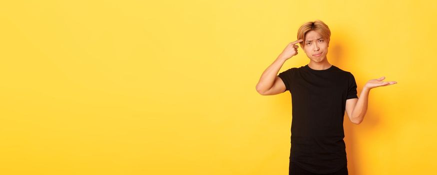 Disappointed asian guy with blond hair, raising hand up puzzled and scolding someone acting stupid, standing yellow background.