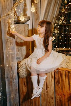 A girl in a festive light dress next to a Christmas tree, lights of garlands in the background. The concept of New Year holidays.