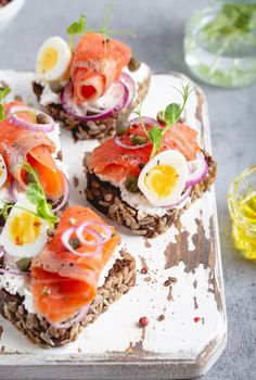 Close-up of sandwich with dark rye bread, cream cheese, salmon, onion, capers, boiled egg on white wooden cutting board, concrete background. Traditional Scandinavian open sandwich Smorrebrod.