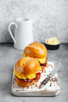 Breakfast sandwiches with scrambled egg, bacon, cheese, tomato on white wooden board, glass with fresh orange juice, white background. Making breakfast concept, selective focus