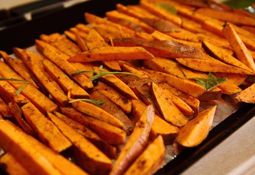 Sliced wedges of organic sweet potato, drizzled with olive oil and seasoned with fragrant culinary herbs and rosemary leaves, on a baking sheet before baking in the oven. Healthy vegan food