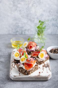 Close-up of sandwich with dark rye bread, cream cheese, salmon, onion, capers, boiled egg on white wooden cutting board, concrete background. Traditional Scandinavian open sandwich Smorrebrod.