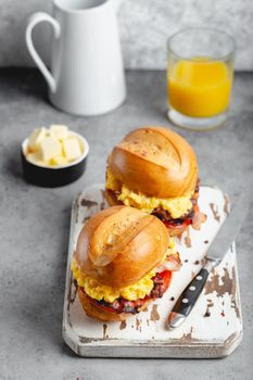 Breakfast sandwiches with scrambled egg, bacon, cheese, tomato on white wooden board, glass with fresh orange juice, white background. Making breakfast concept, selective focus