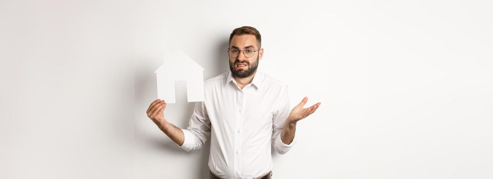 Real estate. Confused man shrugging, showing house paper model and looking indecisive, standing over white background. Copy space