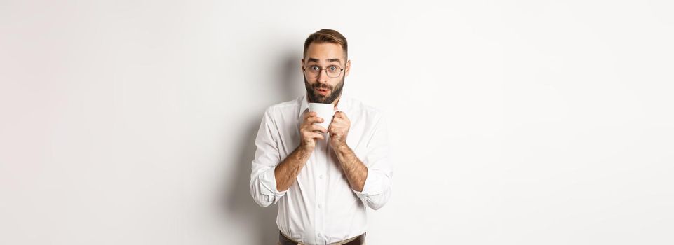 Happy office worker drinking hot coffee and looking excited, gossiping, standing over white background.