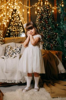A girl in a festive light dress next to a Christmas tree, lights of garlands in the background. The concept of New Year holidays.