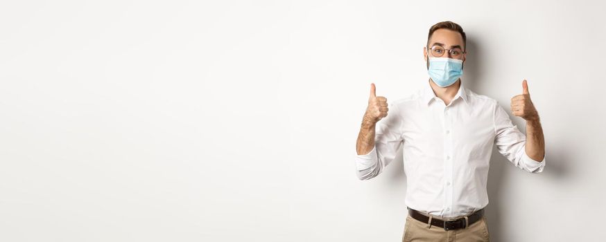 Covid-19, social distancing and quarantine concept. Satisfied male manager showing thumbs up, recommending to wear face mask, standing over white background.