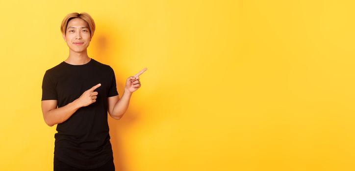 Smiling asian male student with blond hair, pointing fingers right, showing logo, standing yellow background.