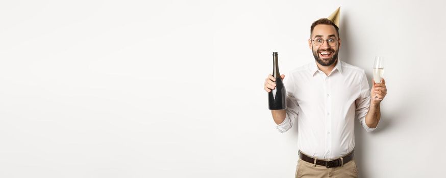 Celebration and holidays. Excited man enjoying birthday party, wearing b-day hat and drinking champagne, standing over white background.