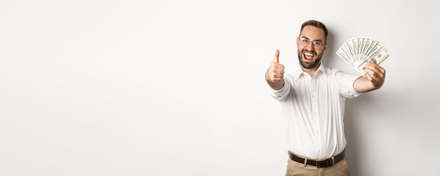 Excited man showing thumbs up and money, earning cash, standing over white background.