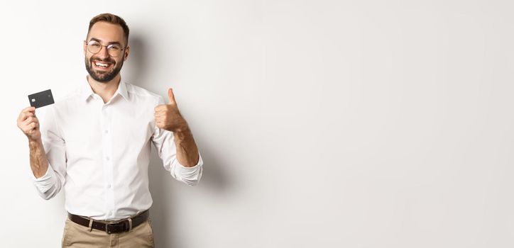 Handsome man showing his credit card and thumb up, recommending bank, standing over white background. Copy space