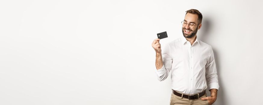 Handsome businessman showing his credit card, looking satisfied, standing over white background.