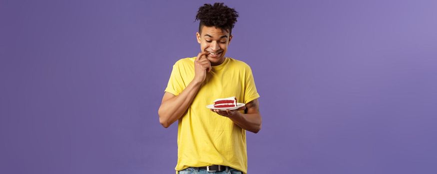 Celebration, party and holidays concept. Portrait of boyfriend cant resist temptation to eat last piece of cake, biting lips and smiling eager to have bite of dessert, hesitating, purple background.