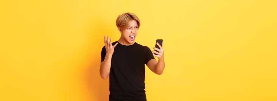 Portrait of angry and pissed-off asian man looking mad at smartphone screen, having argument during video call, standing yellow background.