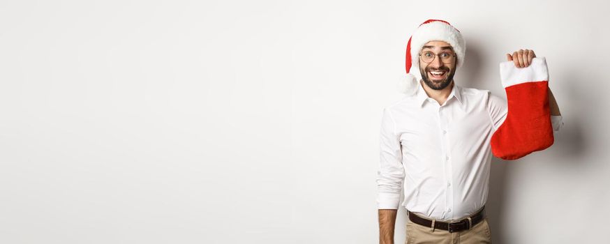 Merry christmas, holidays concept. Excited bearded guy in santa hat holding xmas sock and smiling, celebrating New Year, white background.