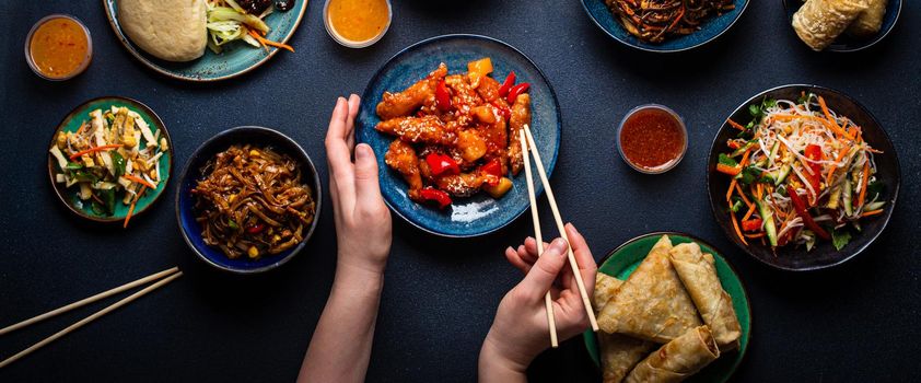 Set of Chinese dishes on table, female hands holding chopsticks: sweet and sour chicken, fried spring rolls, noodles, rice, steamed buns with bbq glazed pork, Asian style banquet or buffet, top view .