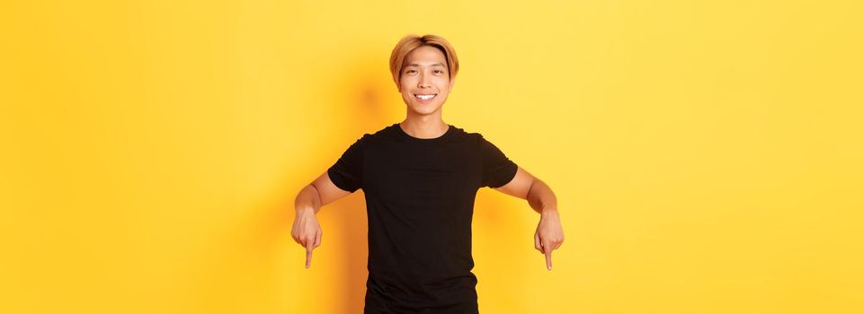 Portrait of smiling attractive korean guy with blond hair, wearing black outfit, pointing fingers down, showing way, standing yellow background.