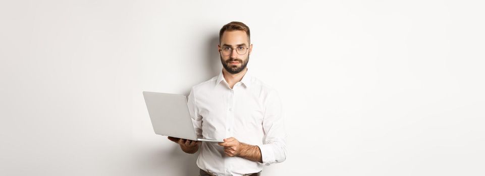 Business. Handsome manager working on laptop, holding computer and looking at camera, standing over white background.