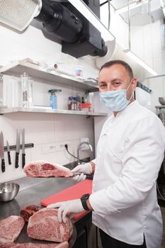 Vertical shot of a chef wearing medical face mask, preparing meat at his kitchen