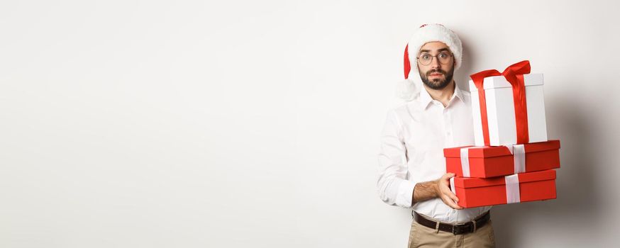 Merry christmas, holidays concept. Confused man in Santa hat holding pile of presents, found gifts under xmas tree, standing against white background.