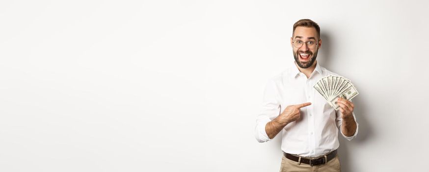 Excited businessman pointing at money, showing dollars and smiling, standing over white background.
