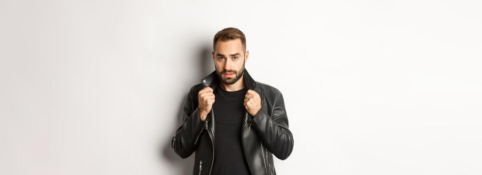 Image of handsome and confident man putting on leather biker jacket, standing against white background.