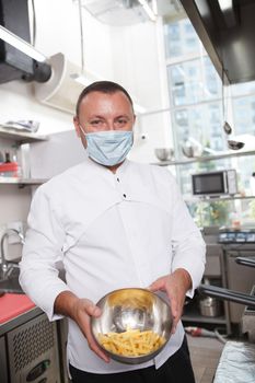 Vertical shot of a professional chef wearing medical face msk, holding a bowl with french fries