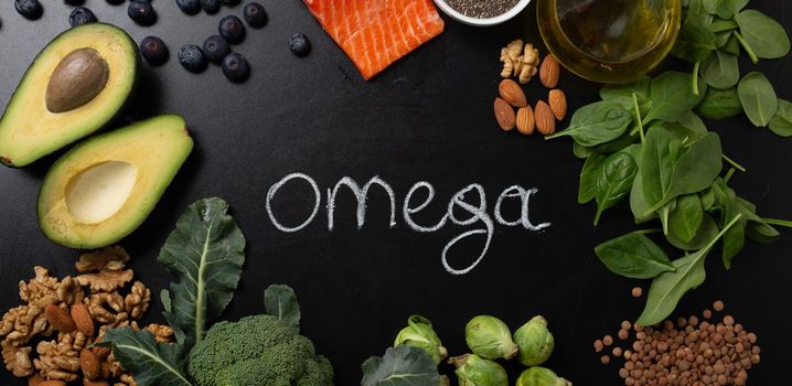 Healthy food with good fat sources, ingredients rich in Omega fatty acids: salmon, vegetables, berries, nuts, seeds, olive oil, black chalk board top view, hand chalk written omega in center .