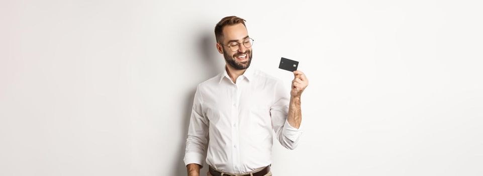 Handsome businessman showing his credit card, looking satisfied, standing over white background.