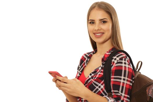 Young beautiful female student smiling happily wearing backpack using her smart phone isolated copy space. Gorgeous woman sending messages. Happy college learner using her cell