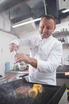 Vertical shot of a cheerful male chef smiling, salting beef on a grill