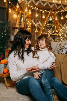 A little girl with her mother in a cozy home environment on the sofa next to the Christmas tree. The theme of New Year holidays and festive interior with garlands and light bulbs.