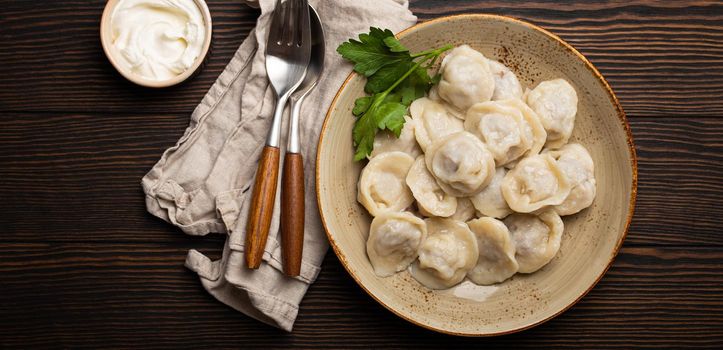 Pelmeni, traditional dish of Russian cuisine, boiled dumplings with minced meat filling on plate with sour cream sauce on wooden rustic background table from above food composition