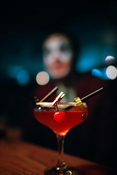 Red cocktail in a glass with a decoration in the form of a marmalade worm and a cherry. Alcoholic drink on the table. Man in clown makeup on the background. Halloween party at a nightclub.