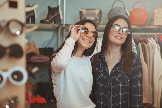 Two lovely cheerful female friends trying on sunglasses, while shopping together at clothing store. Beautiful women choosing eyewear at accessories shop