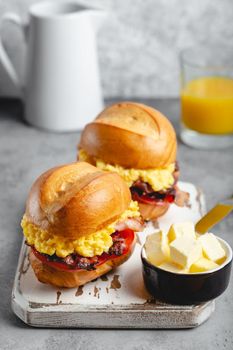 Breakfast sandwiches with scrambled egg, bacon, cheese, tomato on white wooden board, glass with fresh orange juice, white background. Making breakfast concept, selective focus