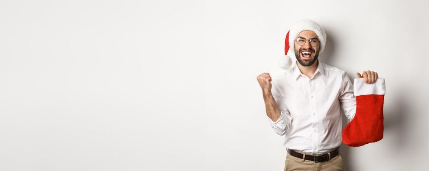 Merry christmas, holidays concept. Happy adult man receive gifts in xmas sock, looking excited, wearing santa hat, white background.