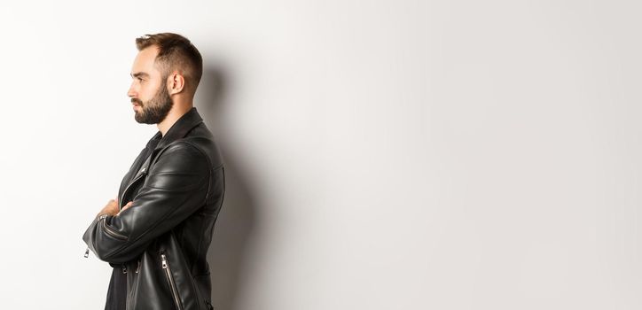 Profile of handsome serious man in leather jacket, looking left, holding hands on chest confident, white background.