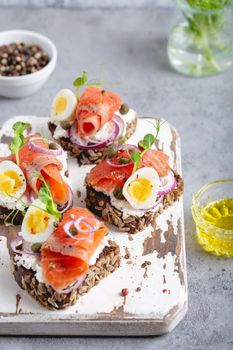 Close-up of sandwich with dark rye bread, cream cheese, salmon, onion, capers, boiled egg on white wooden cutting board, concrete background. Traditional Scandinavian open sandwich Smorrebrod.