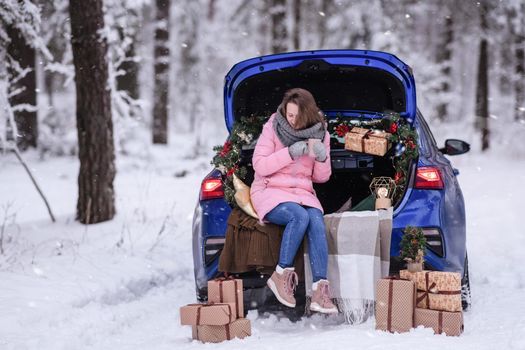 A woman in winter clothes drinks a hot drink sitting in a car decorated in a New Year's way. Traveling by car through the forest. A trip before Christmas.