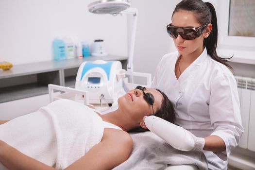 Professional cosmetologist using laser hair removal machine, working with female client. Young woman getting laser skincare treatment at beauty clinic