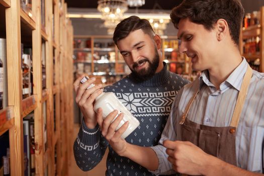 CHeerful sommelier helping his male customer choosing alcohol for the party. Bearded man buying whiskey at the supermarket