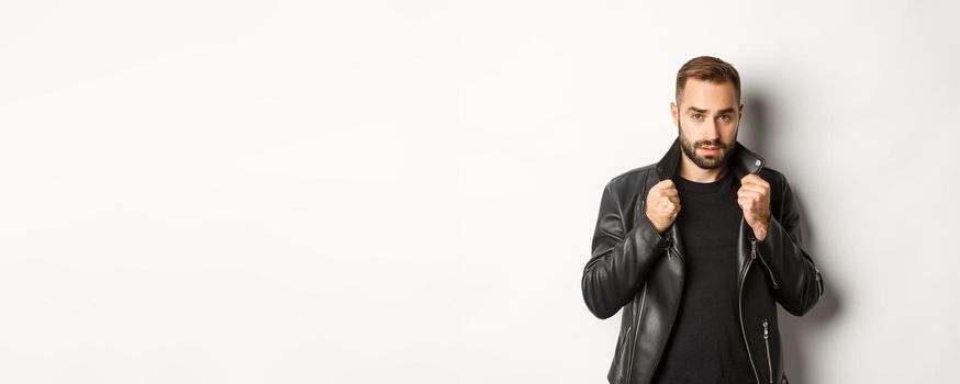 Image of handsome and confident man putting on leather biker jacket, standing against white background.