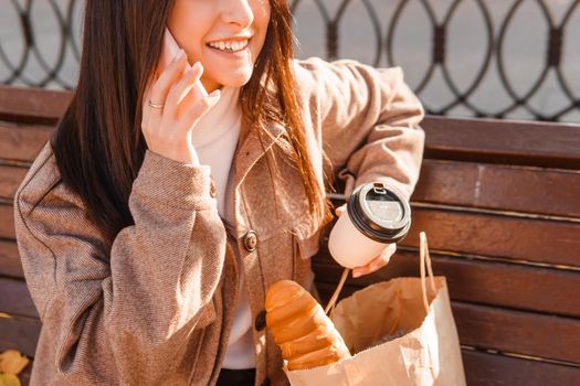 A stylish brunette woman walks around the autumn city. The brunette is sitting on a bench outside with a glass of coffee and a phone, holding a craft bag with a baguette.