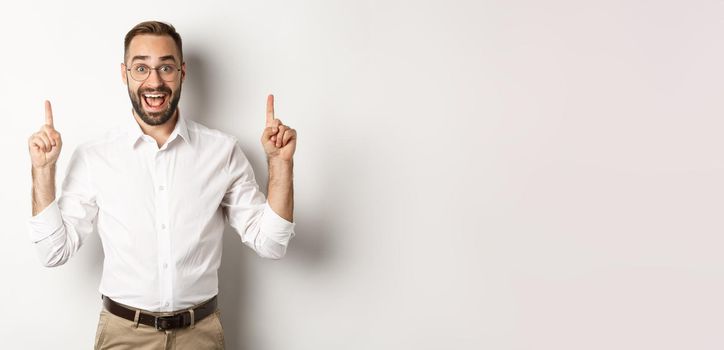 Happy male entrepreneur in business clothes, pointing fingers up and showing promo offer, standing over white background.